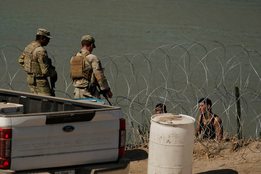 Two men in army uniforms speak with people below standing in the water behind a wire fence.