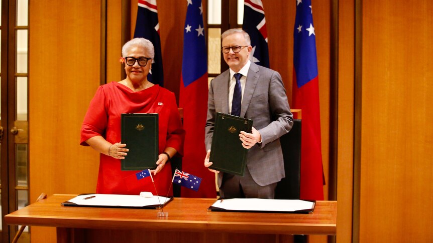 A woman and a man display leatherbound formal documents as they stand before Samoan and Australian flags.