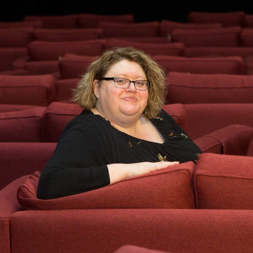A woman with chin-length dark blond hair, wearing a black top and glasses, sitting in a theatre with red seats.