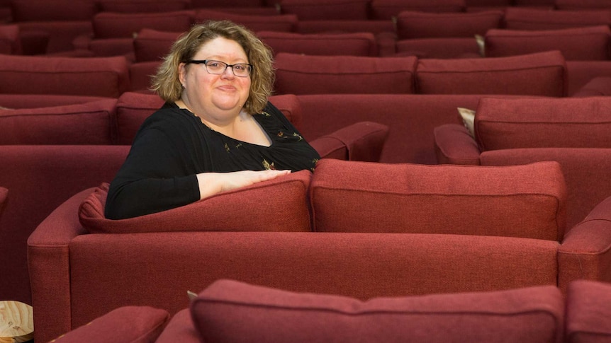 A woman with chin-length dark blond hair, wearing a black top and glasses, sitting in a theatre with red seats.