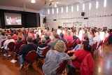 A crowd of people sit in a hall and watch a large screen.