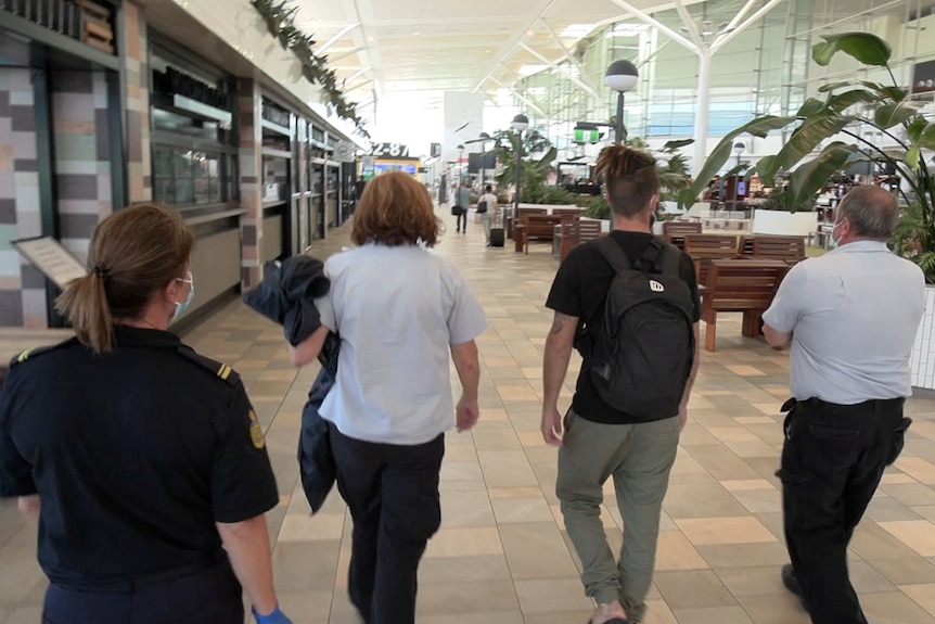 A man wearing a black t-shirt and olive trousers is flanked by security guards.