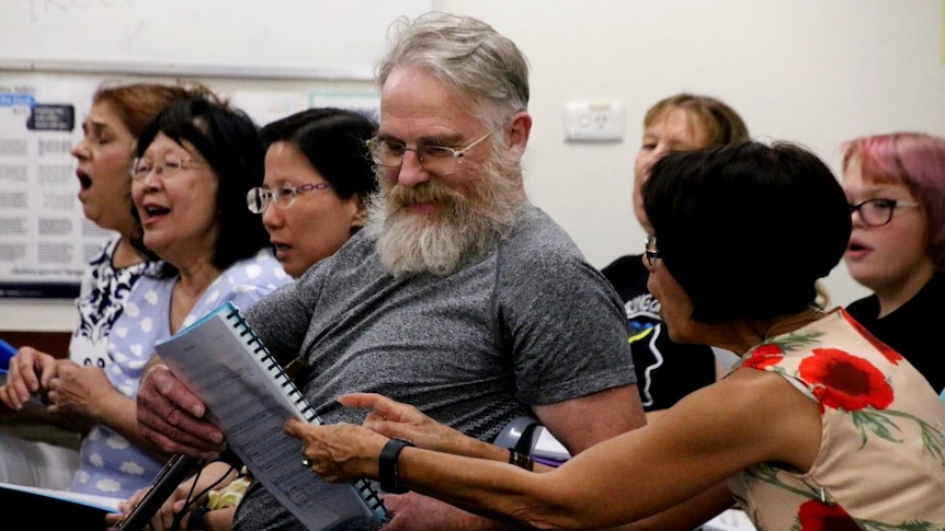 A man with a grey t-shirt and glasses looks at sheet music handed to him by a woman wearing a floral dress.