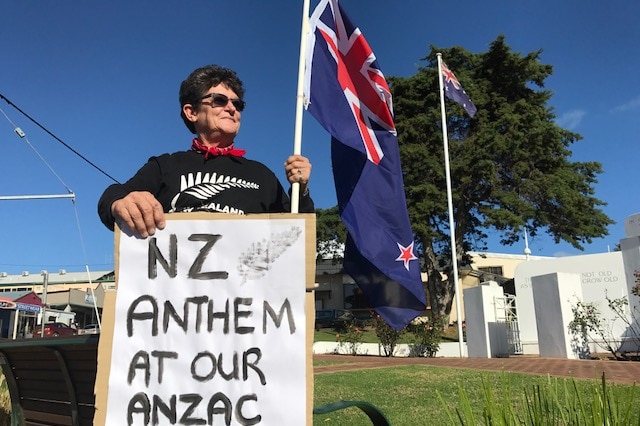 A woman holding a sign and New Zealand flag standing in front of a war memorial