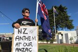 A woman holding a sign and New Zealand flag standing in front of a war memorial