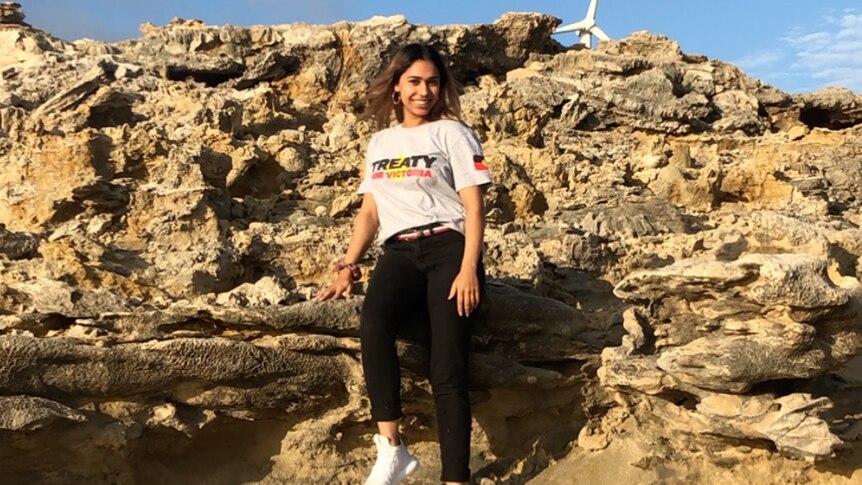 Shahnaz Rind, wearing a white shirt that says 'treaty' standing on rocky cliffs. 