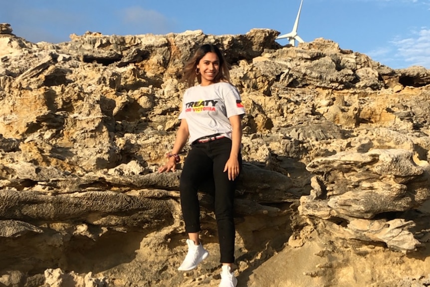 Shahnaz Rind, wearing a white shirt that says 'treaty' standing on rocky cliffs. 