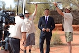 Stan Grant holds a microphone and smiles as a tv crew works around him.