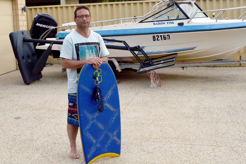 Paul Goff stands in front of a boat holding a blue bodyboard.