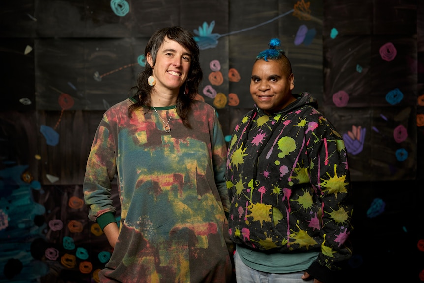 First Nation Australian (right) and first generation Australian with English heritage (left) smiling in front of their artwork.