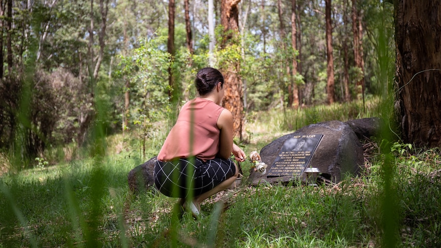 Woman kneels in front of headstone in bushland cemetery, pictured in story about eco-friendly funerals.