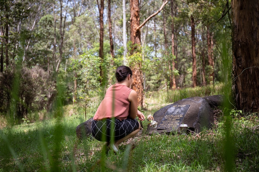 Woman kneels in front of headstone in bushland cemetery, pictured in story about eco-friendly funerals.