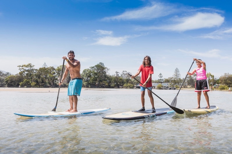 Three people on stand-up paddle boards taking part in an Aboriginal cultural tour on Coffs Creek, Coffs Harbour NSW.