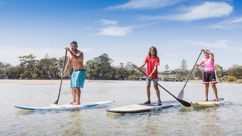Three people on stand-up paddle boards taking part in an Aboriginal cultural tour on Coffs Creek, Coffs Harbour NSW.