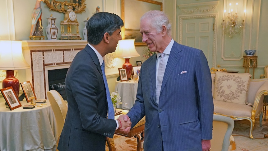 An older white man with white hair in a navy pinstriped suit smiles as he shakes hands with a middle-aged man of Indian heritage
