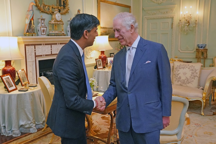 An older white man with white hair in a navy pinstriped suit smiles as he shakes hands with a middle-aged man of Indian heritage