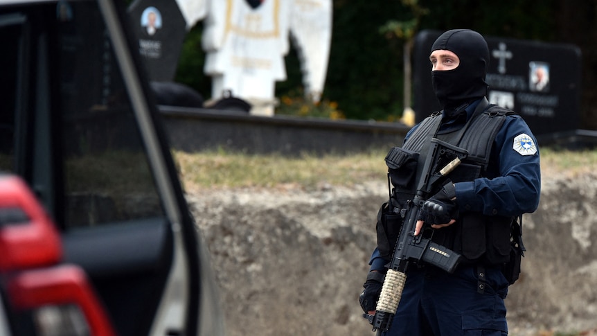 A police officer in a balaclava holding a rifle patrols a road in front of a white angel statue.