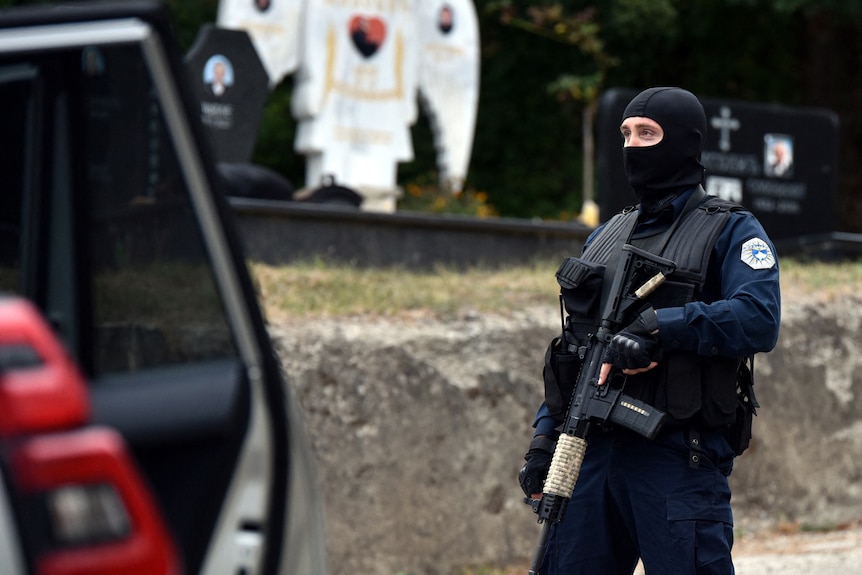 A police officer in a balaclava holding a rifle patrols a road in front of a white angel statue.