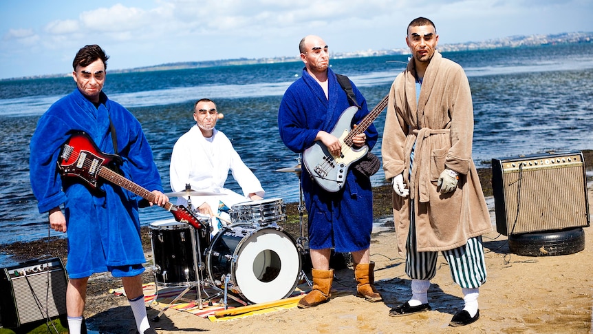 Four members of Eddy Current Suppression Ring performing by the water. They wear robes and masks.