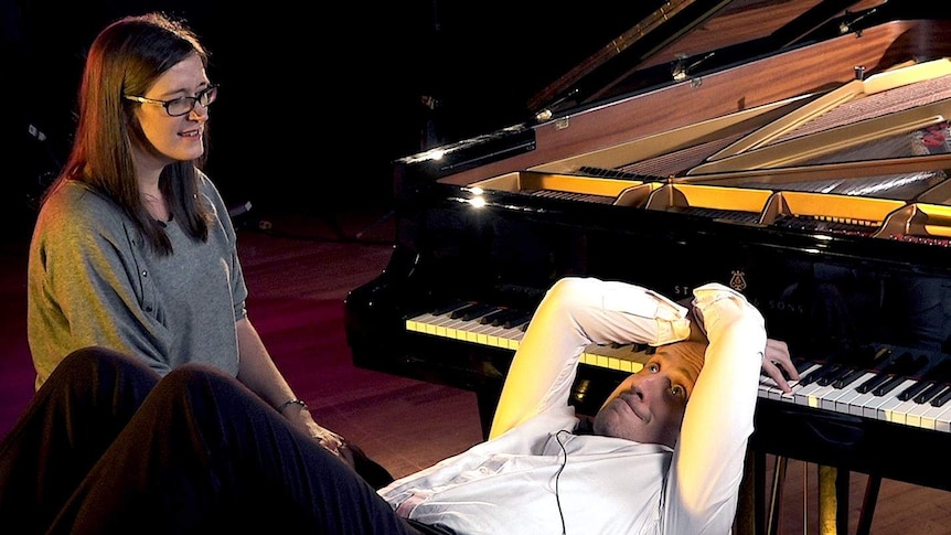 Dr Jennifer MacRitchie sitting next to pianist Simon Tedeschi as he playe the piano lying on his back on the piano stool.