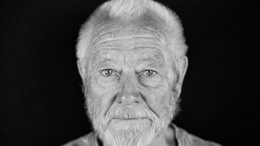 A black and white photo of a man which white hair and a white beard, wrinkled forehead looking forward at camera