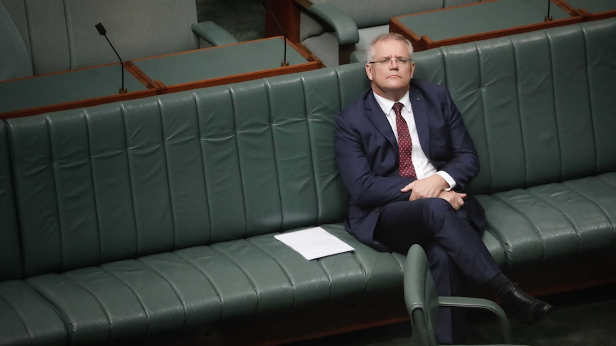 Morrison is sitting on the front bench by himself, looking upwards in a reflective pose.