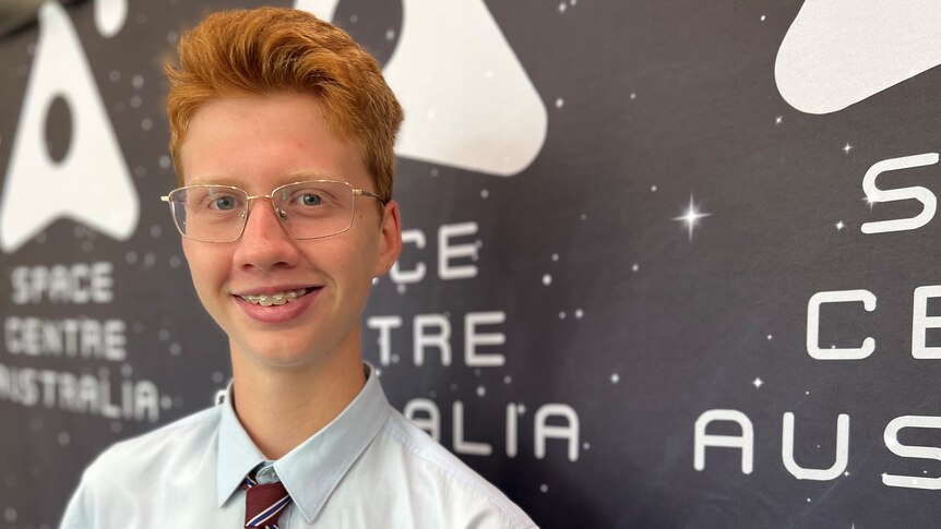 A smiling teenager with ginger hair and glasses, wearing a school uniform.