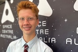 A smiling teenager with ginger hair and glasses, wearing a school uniform.