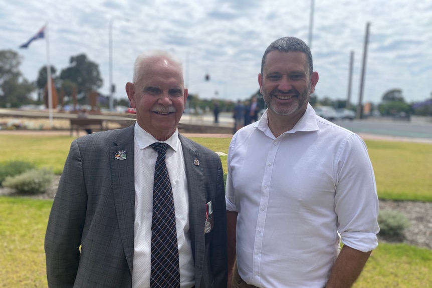 Two Aboriginal men, one dressed in a suit, the other in pants and white shirt smile for a photo at an outdoor location