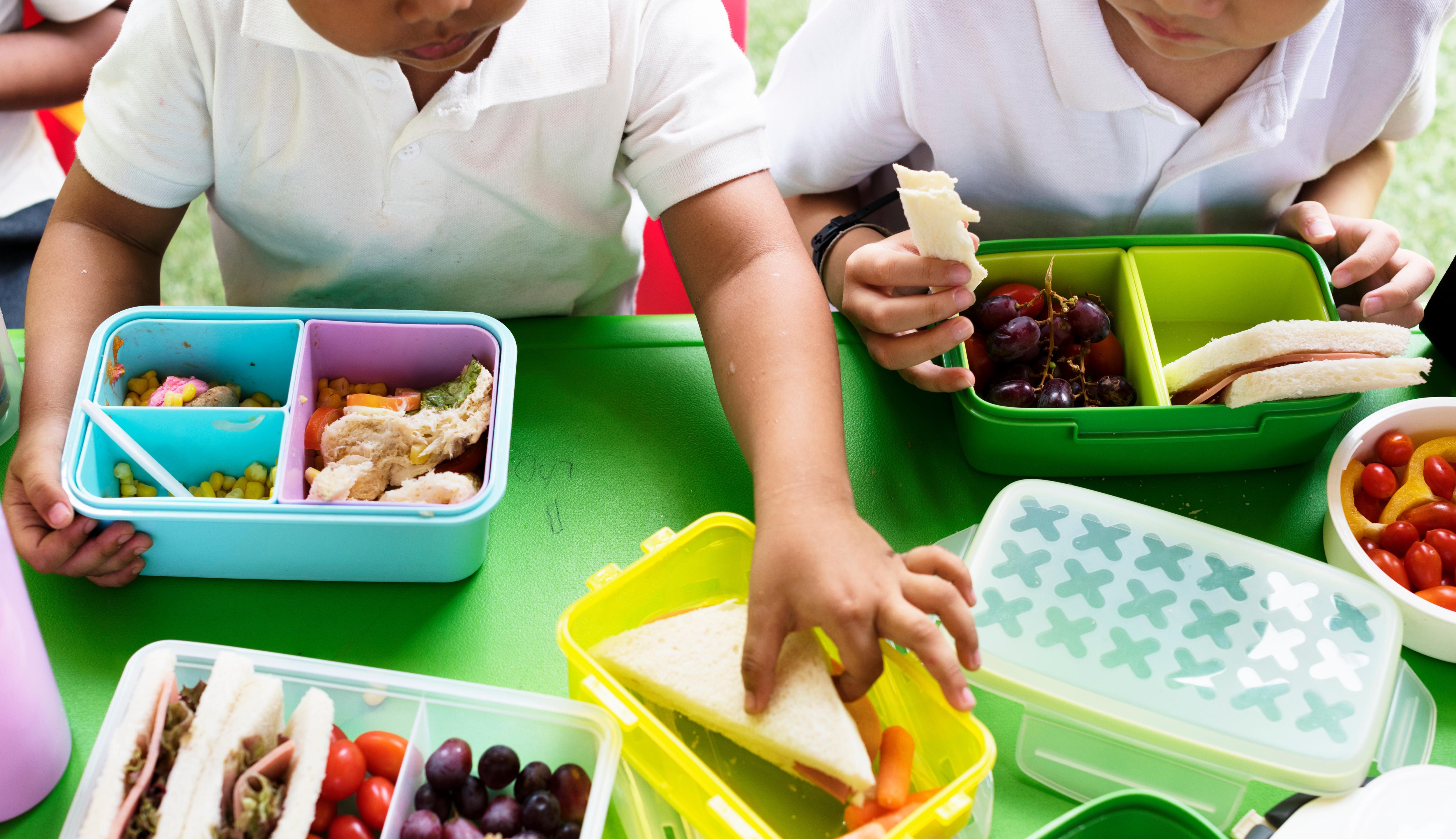 Two young children reach for sandwiches, grapes and carrots from colourful lunch boxes at school.