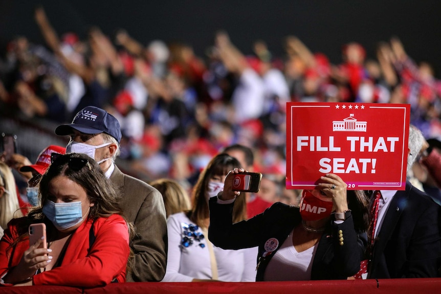 A woman holding a sign reading 'Fill That Seat!' at a Trump rally