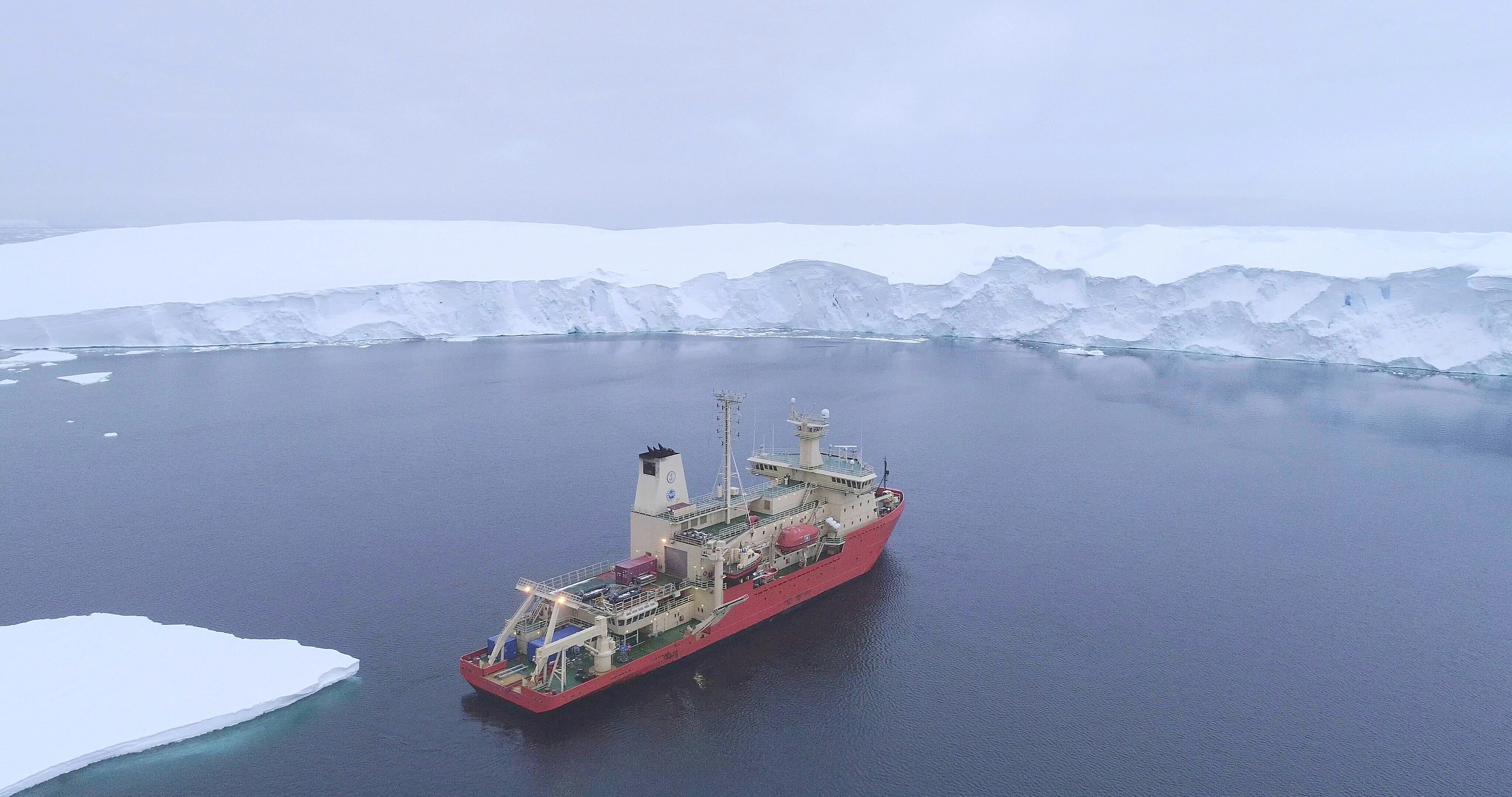 A red scientific research ship floats in blue water in front of a large white glacier.