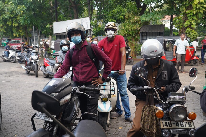 A group of men stand with motorcycles in a queue on a paved street