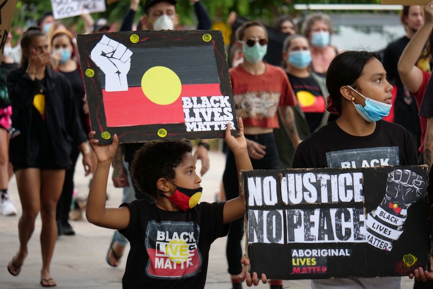 Walali holds up a Black Lives Matter sign, wearing an Aboriginal flag mask and Black Lives Matter shirt, marching in crowd.