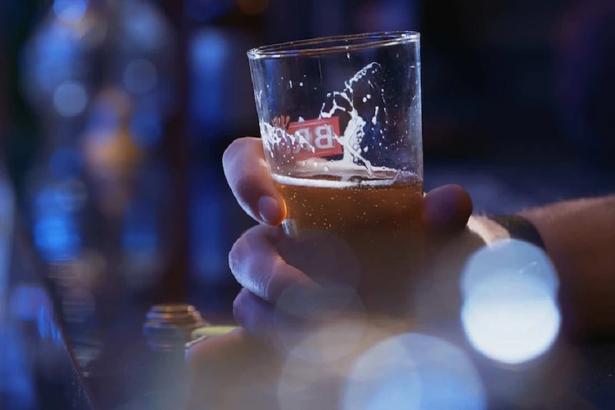 A hand holding a glass half-filled with beer with a blurred background suggesting a bar or pub setting.