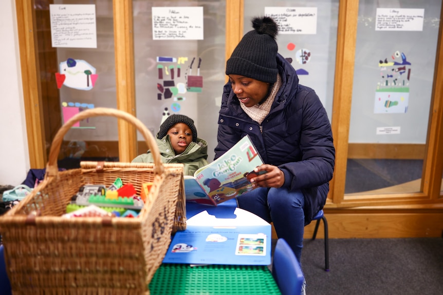 A woman wearing a puffer jacket and beanie reads a book to a little child.