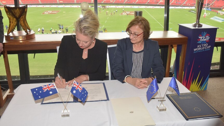 Two women sit at a desk signing a piece of paper with a pen, with a stadium in the background.