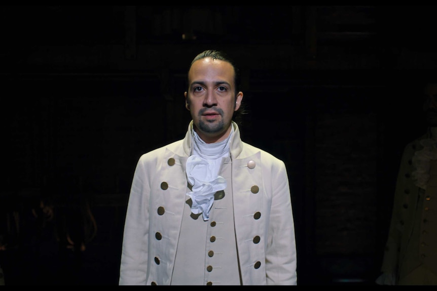 Lin-Manuel Miranda in a scene from Hamilton the musical, he stands in the middle of the stage in a white outfit