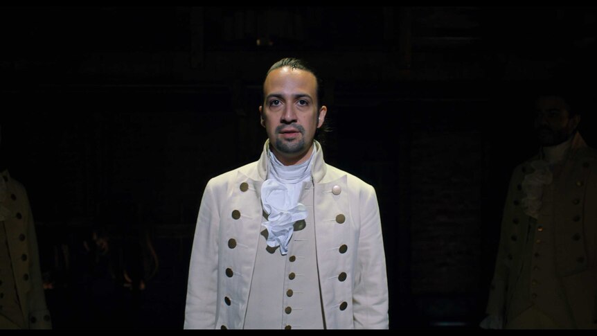 Lin-Manuel Miranda in a scene from Hamilton the musical, he stands in the middle of the stage in a white outfit