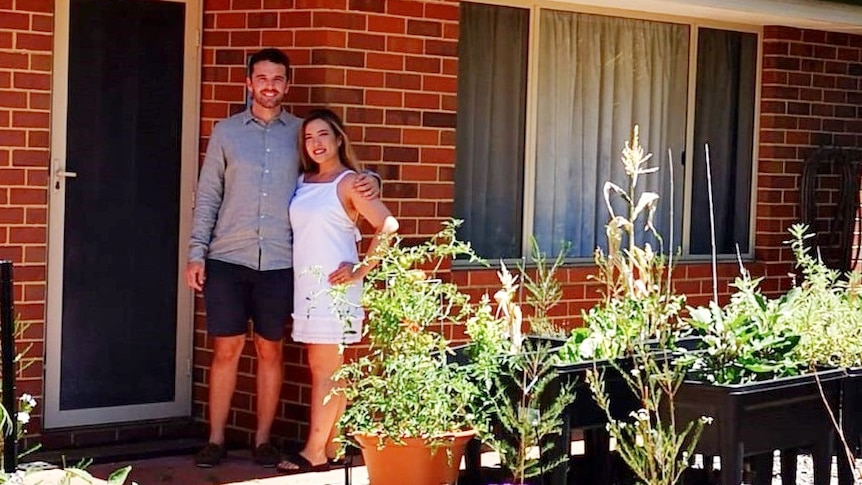 A young couple smile in front of a red brick house