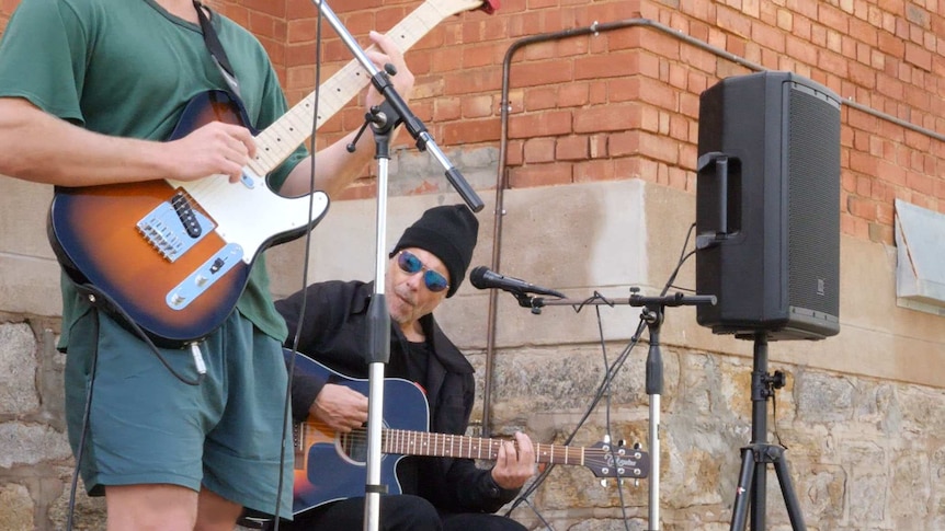 A man in green clothing plays an electric guitar, accompanied by a man in black playing an acoustic guitar.