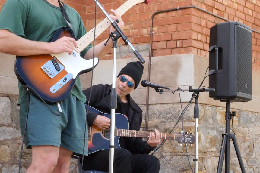 A man in green clothing plays an electric guitar, accompanied by a man in black playing an acoustic guitar.