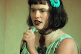 Eurydice Dixon stands and speaks into a microphone, wearing a green-tinged dress.