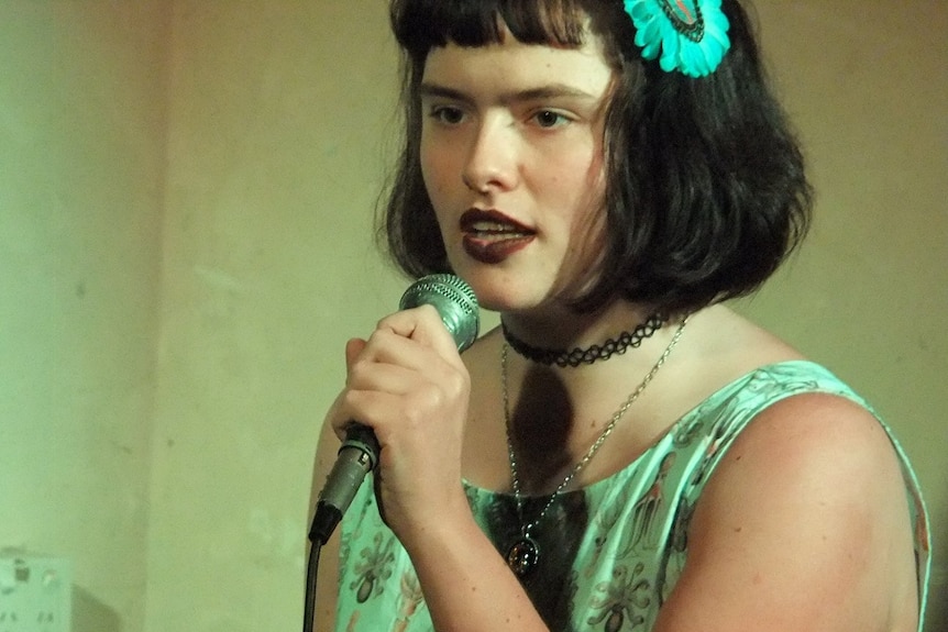 Eurydice Dixon stands and speaks into a microphone, wearing a green-tinged dress.