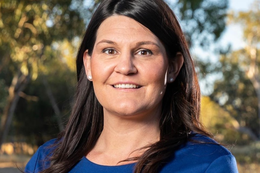 A woman smiles at the camera. She has long dark hair and is wearing a blue long sleeved top.