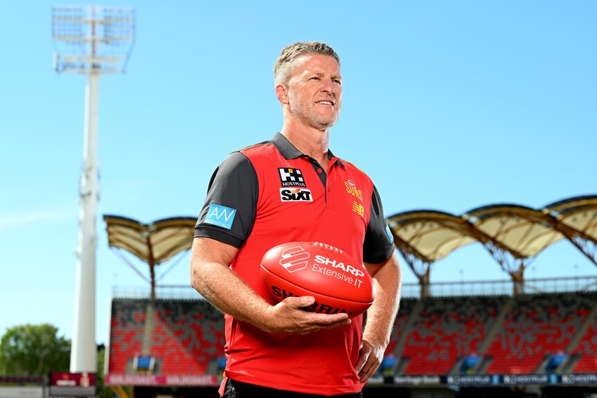 Damien Hardwick stands holding a footy while wearing a Gold Coast Suns polo