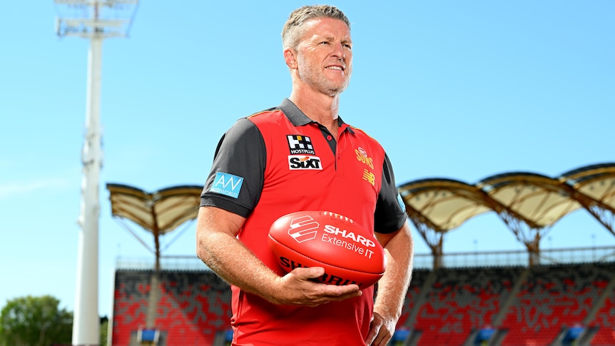 Damien Hardwick stands holding a footy while wearing a Gold Coast Suns polo