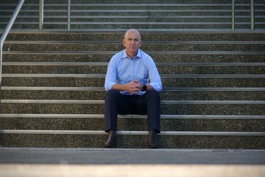 A bald man in a blue business shirt sitting on stairs.