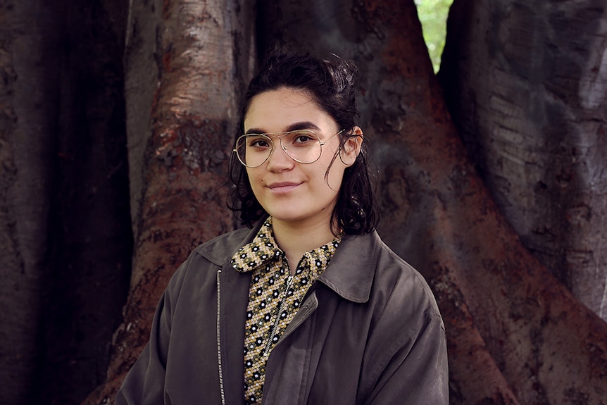 A girl with dark hair and glasses stands outdoors in front of the base of a large tree in the shade.