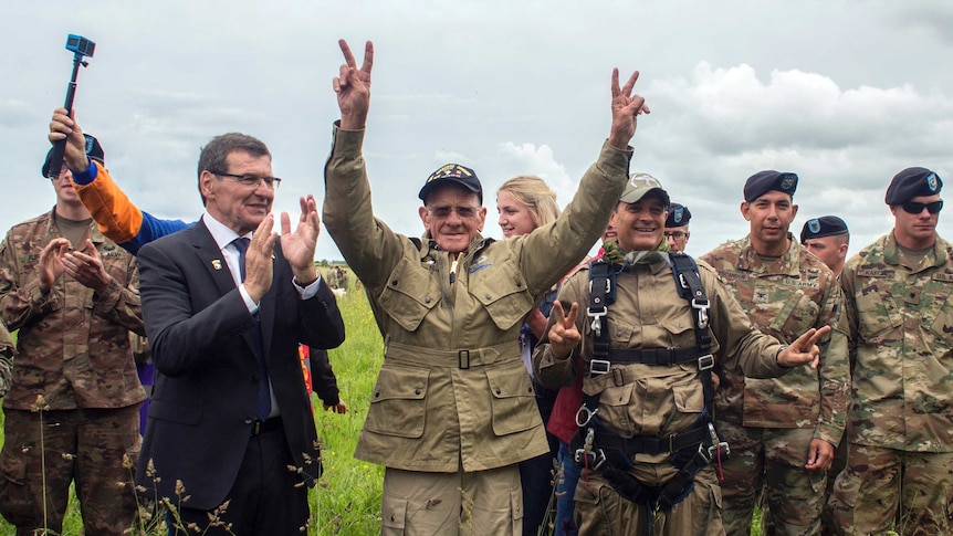 97yo in army fatigues holds two victory signs in the air surrounded by other parachutists in a field.